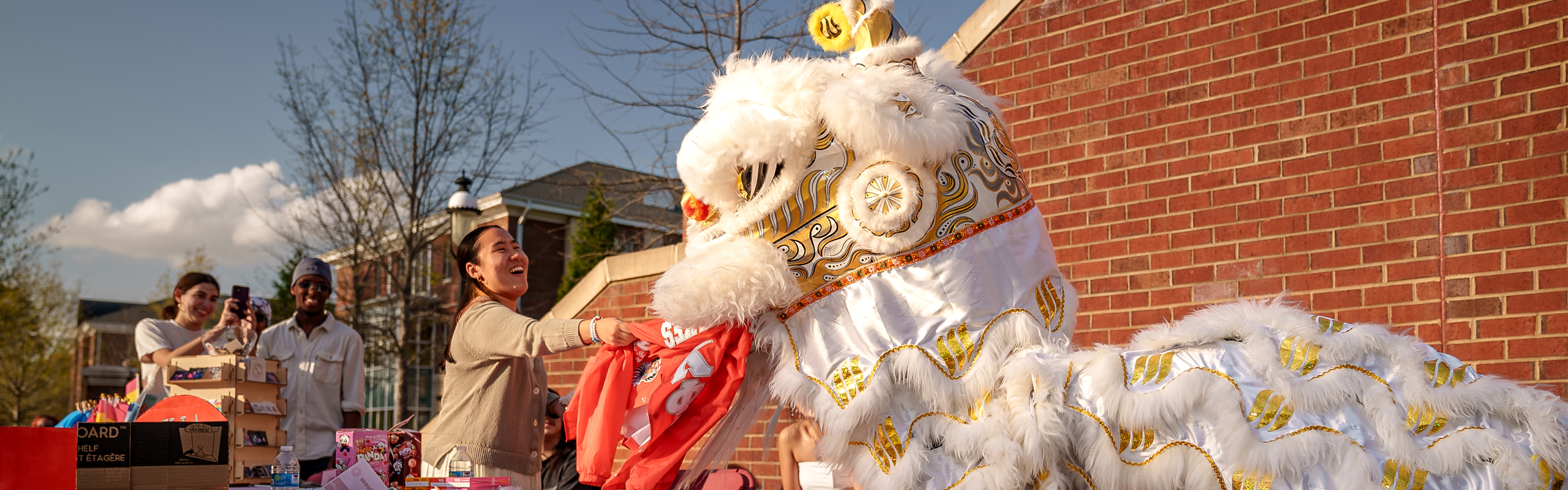 A student interacts with a traditional Chinese lion dance costume during the Asian and Pacific Islander Heritage Month Kickoff at Elon University, surrounded by colorful decorations and smiling onlookers.