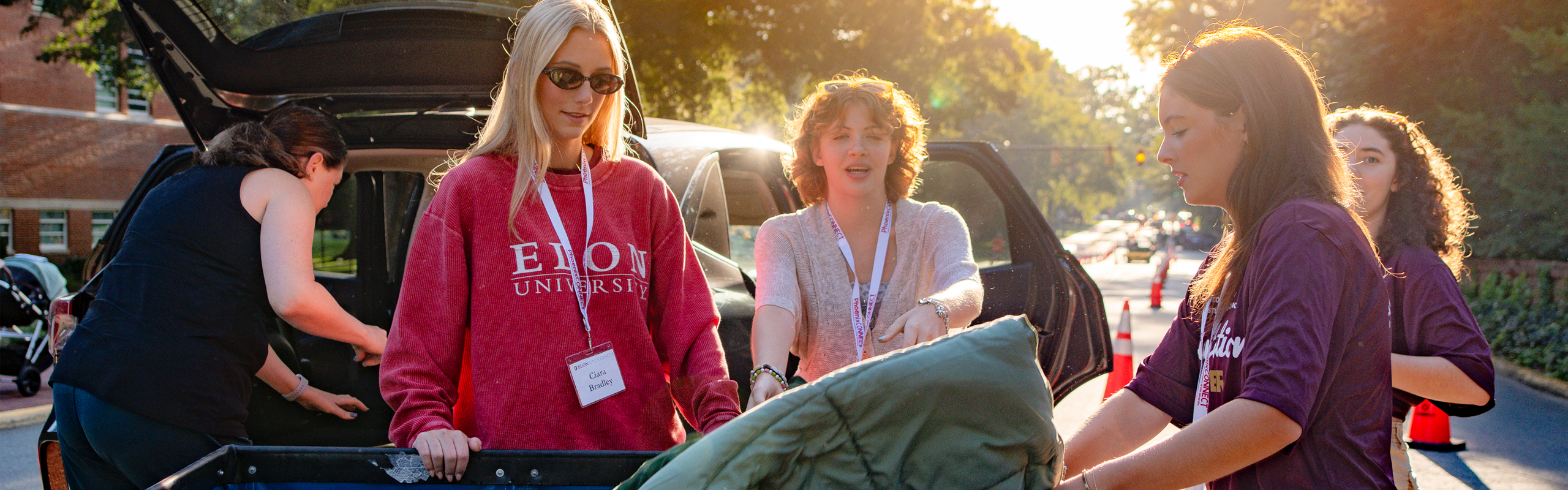 Elon University students help move belongings from a car during early morning move-in, wearing name tags and orientation shirts as sunlight filters through the trees.