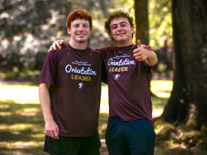 Two Elon University Orientation Leaders smile and pose for a photo during Move-In Day, wearing matching maroon shirts and standing outdoors in a shaded campus area.