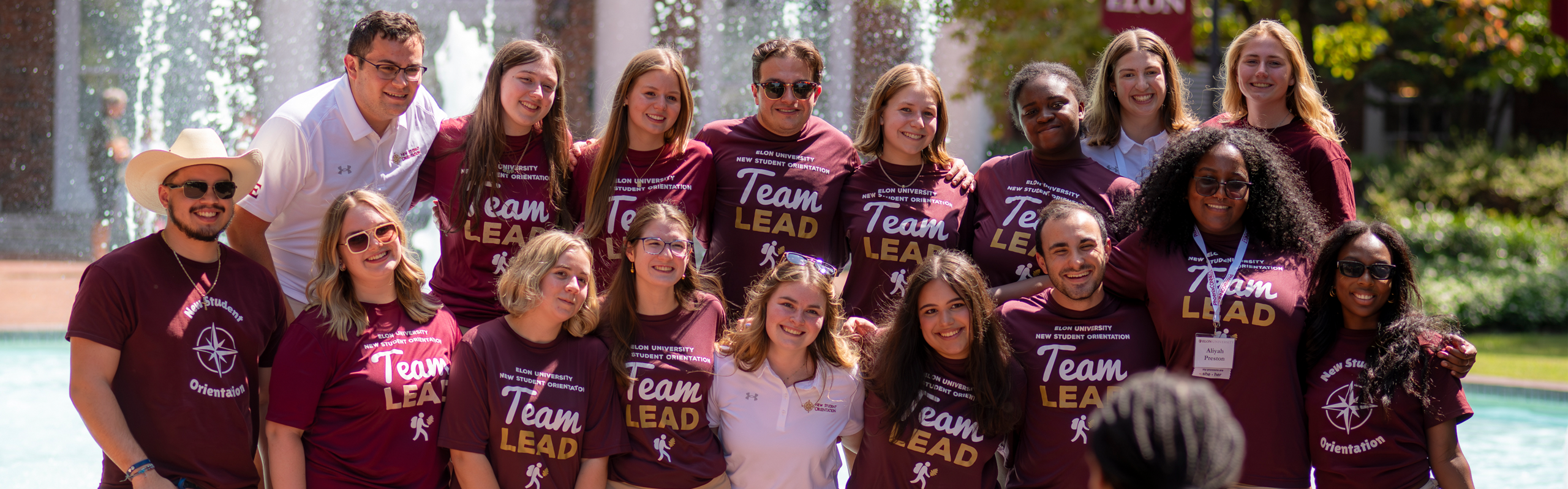 Elon University New Student Orientation leaders smile for a group photo in front of a fountain, wearing matching maroon “Team Lead” shirts and name tags.