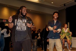 Two individuals dance and smile during the Black History Month Kickoff celebration at Elon University. One person wears a "Black Voters Matter" shirt and holds a camera, while others enjoy the festive atmosphere in the background.