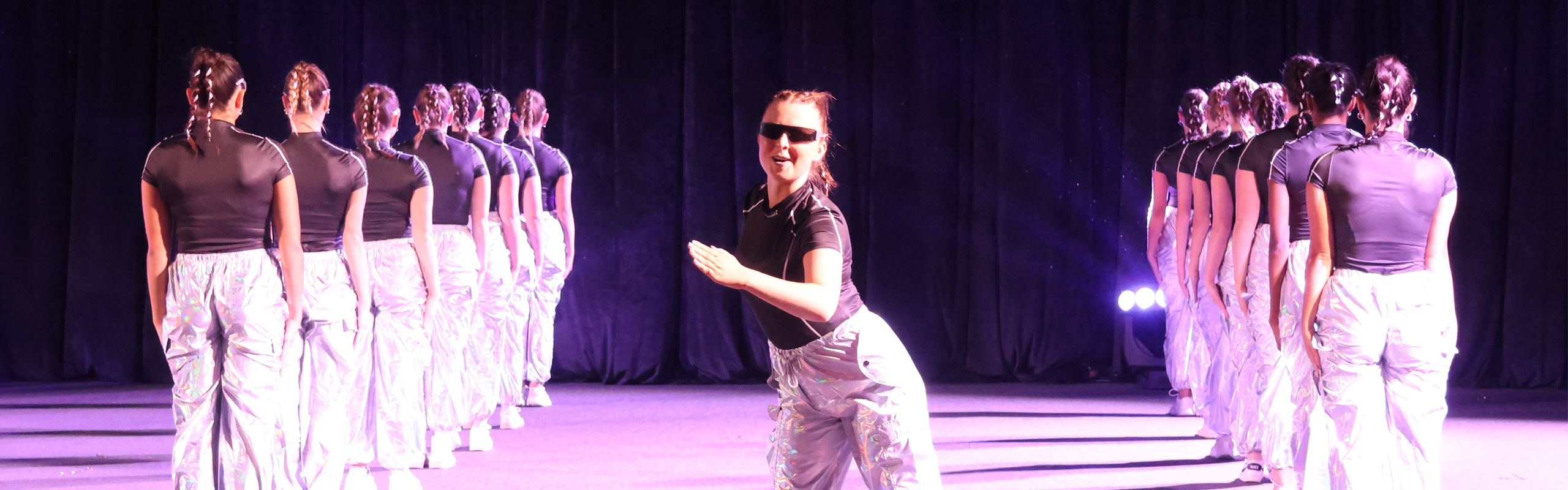 A group of dancers with braided hair perform on stage in matching metallic pants and black tops. One dancer at the front faces the audience while the rest face the back.
