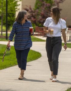 Two women walk side by side on a sunny sidewalk, smiling and holding iced drinks, engaged in conversation, with greenery and campus buildings in the background.