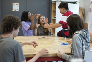 Five students collaborate at a wooden table in a maker space, using spaghetti sticks, tape, and string to build a structure, demonstrating teamwork and creativity in a hands-on activity.