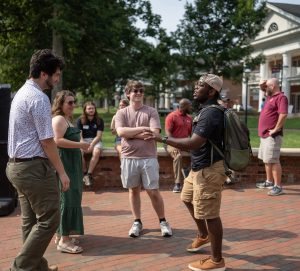 A group of people stand and chat outdoors on a brick pathway during a sunny day at College Coffee. One man with a backpack gestures animatedly while others smile and listen.