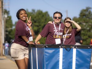 Three student orientation leaders in matching maroon shirts smile while standing by a large blue move-in cart on a sunny day. One flashes a peace sign, another shades their eyes from the sun.