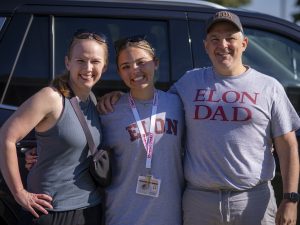 Smiling student stands between two parents on Move-In Day, wearing an Elon shirt and lanyard, with her father in an “Elon Dad” T-shirt and her mother in a tank top and sunglasses.