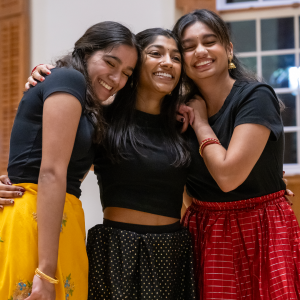 Three students dressed in colorful skirts and black tops share a joyful hug during a Diwali celebration, smiling brightly in a warmly lit room.