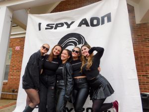 Five students dressed in black pose together in front of a white banner reading “I SPY ΑΟΠ” with a graphic logo, during an Alpha Omicron Pi sorority event at Elon University.