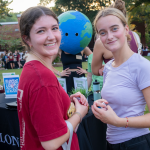 Two students smile at the camera during a student organization fair at Elon University, with event booths, a QR code sign, and a person wearing an Earth costume in the background.