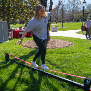 A student balances on a slackline during HealthEU Day, enjoying outdoor activities on a sunny campus lawn surrounded by event booths, trees, and fellow students.