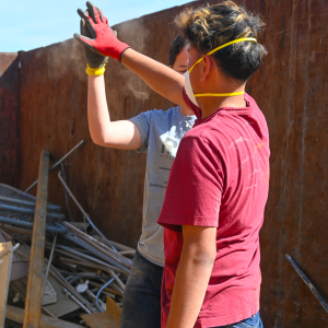 Two students wearing work gloves and dust masks give each other a high five while standing in a large dumpster filled with construction debris and scrap metal during a volunteer clean-up project.