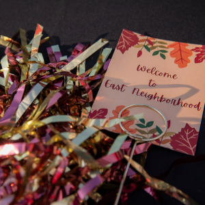A festive sign reading "Welcome to East Neighborhood" is displayed alongside a colorful bunch of metallic maroon, gold, and silver pom-poms on a dark tablecloth.