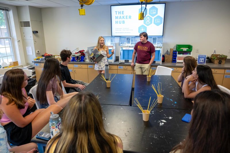 In "THE MAKER HUB" classroom, a student presents to her peers, holding a laptop. Beside her, a student in an "Elon Engineering" shirt observes. Others sit attentively around tables with crafting materials.