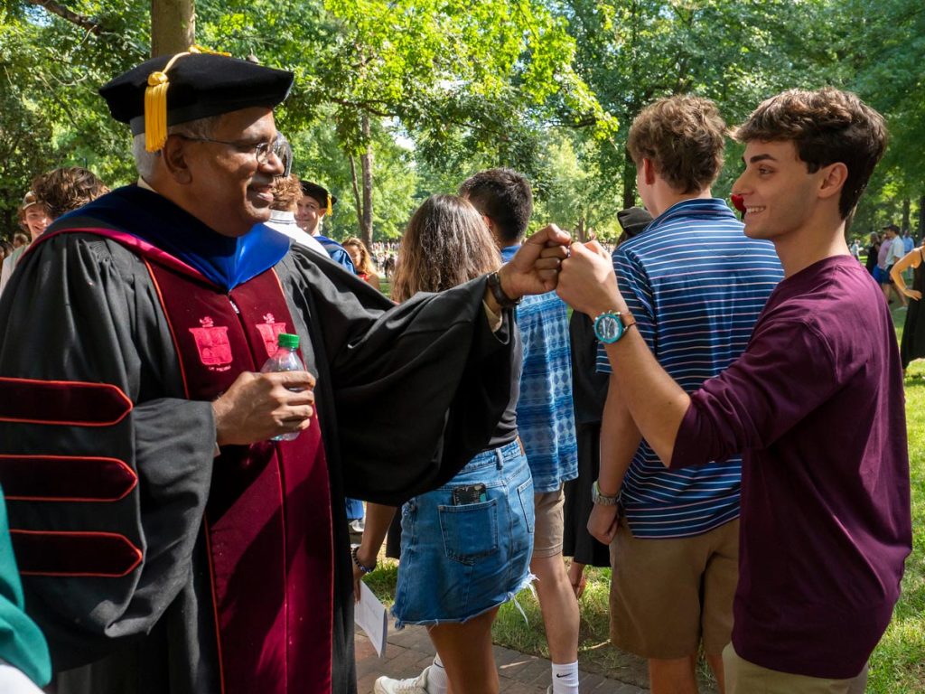 The dean of Elon's business school, dressed in traditional graduation regalia and holding a water bottle, shares a fist bump with a young man in a burgundy shirt. They are at the new student convocation ceremony, surrounded by a diverse crowd with trees as a natural backdrop.
