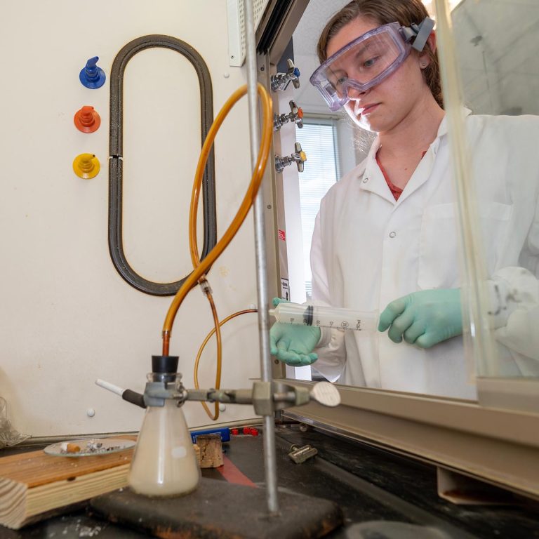 An Elon student, dressed in a lab coat, safety goggles, and gloves, carefully employs a syringe to inject a substance into a long tube connected to a beaker.