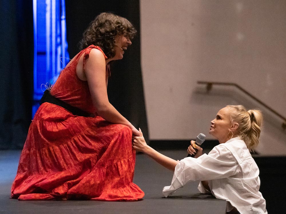 A woman in a red dress kneels on a stage, while actress and singer Kristin Chenoweth stands before the stage in a white shirt, holding a microphone and extending her hand to grasp the hand of the woman in the red dress.