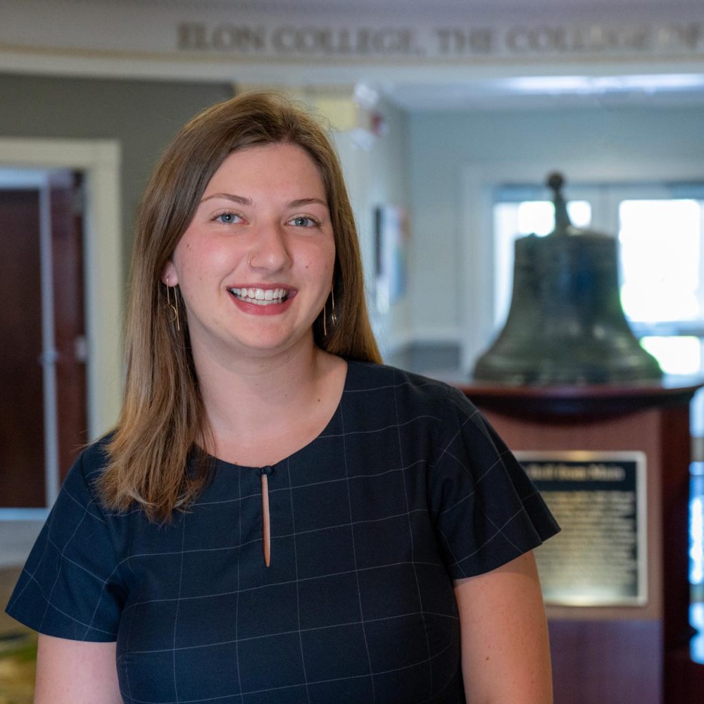 Megan Curling smiles in a navy dress as she stands in front of the historic bell, located in the rotunda of Alamance Building on Elon University's campus.