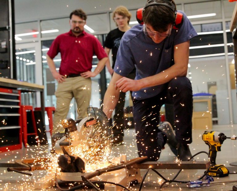 A student using a saw to cut metal, throwing a shower of sparks onto the floor of the prototype lab.