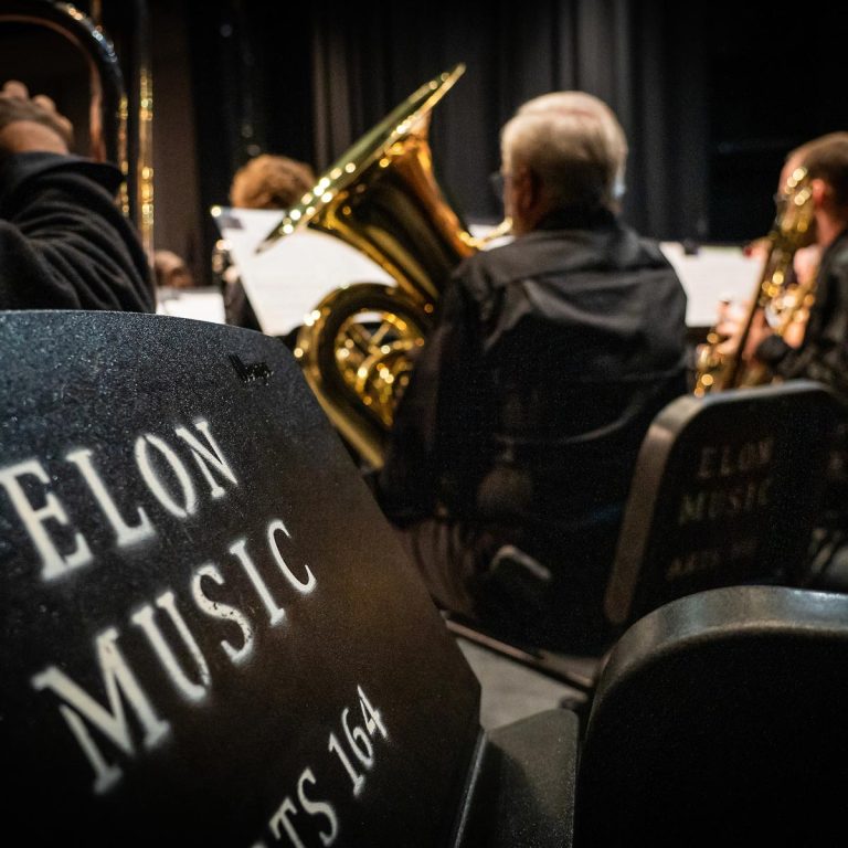 A group of musicians performing with their instruments in a concert hall, with a music stand prominently displaying 'Elon Music' in the foreground.