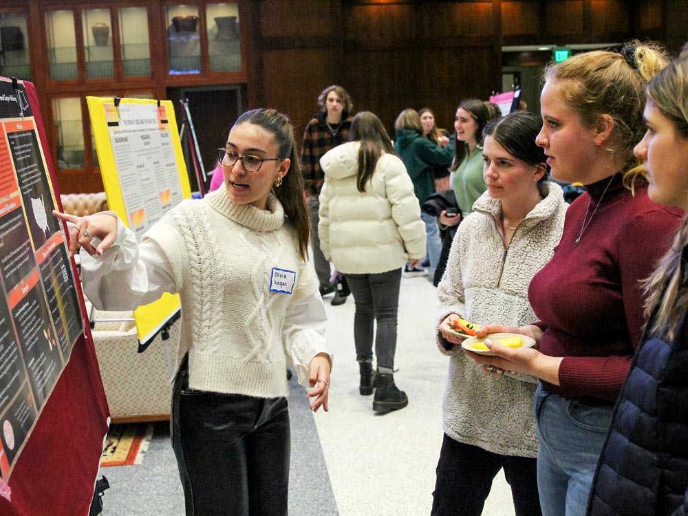 An Elon College junior fellow gestures towards her research poster while engaging in a presentation about her research to a group of attentive individuals.
