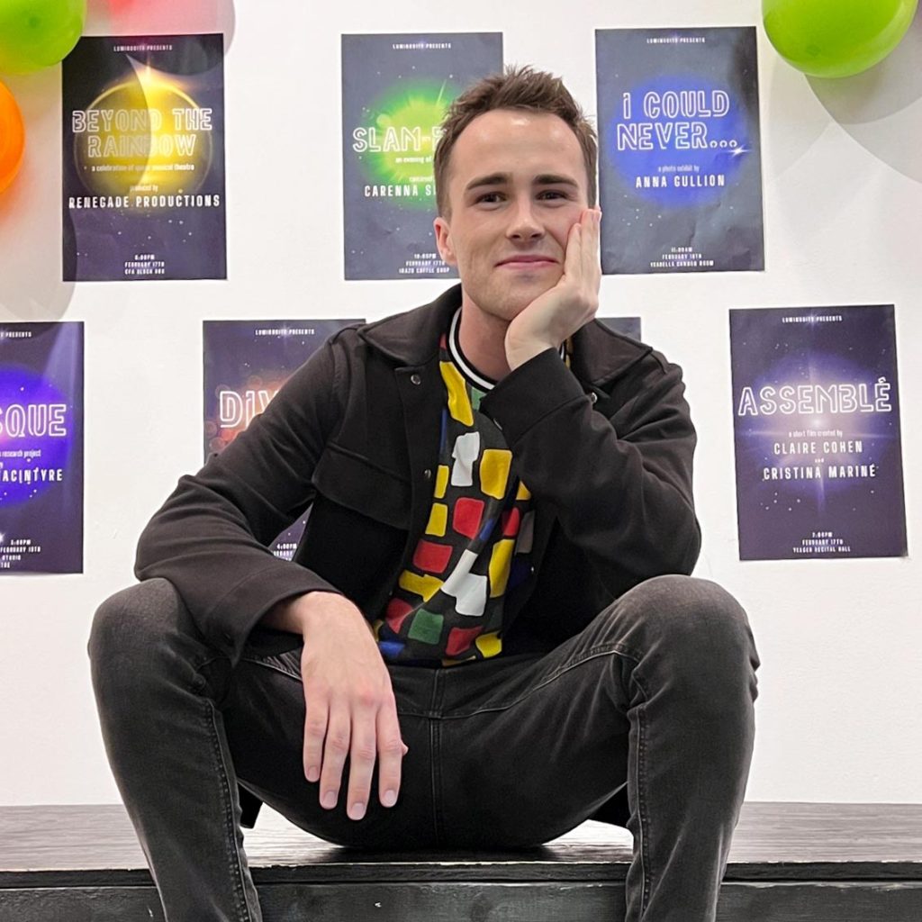 Jack Morrill sitting on the edge of a stage with his chin resting in his palm, in front of a wall covered in balloons and posters.