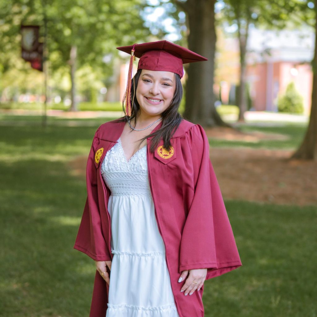 Maddy Starr poses under the oaks on Elon University's campus, dressed in a maroon graduation cap and gown.