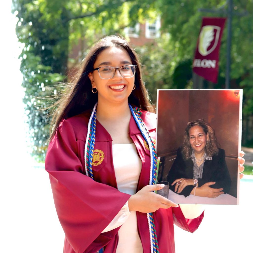 Ons Yasmeen Mrad Bouali stands in front of a campus fountain at Elon University, dressed in a maroon graduation gown and holding a photo of her mother.