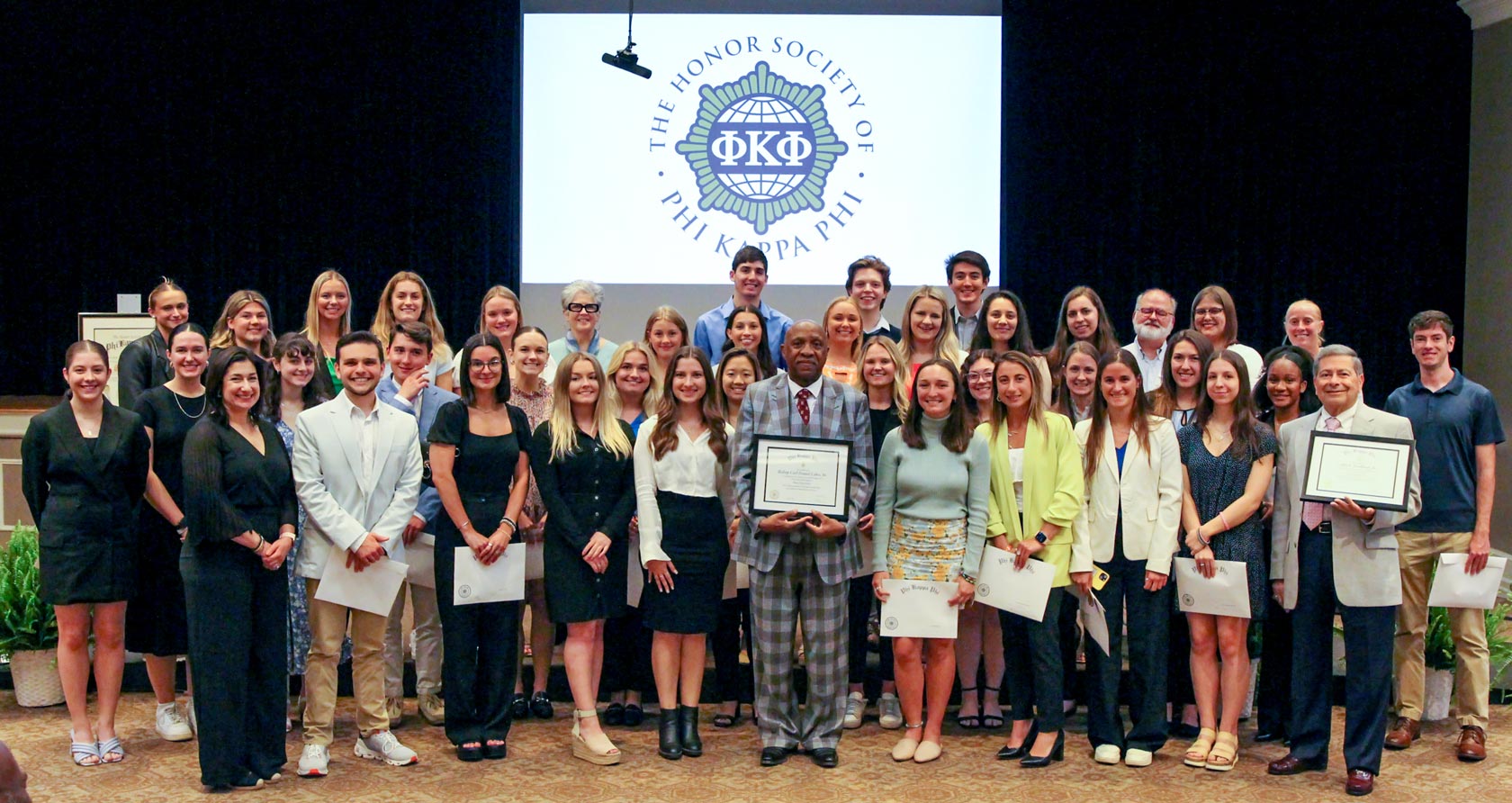A substantial gathering of newly initiated Phi Kappa Phi members stands united on a stage, with the Phi Kappa Phi logo displayed on a background screen. Several members proudly hold certificates.