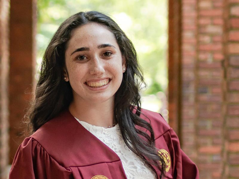 Talya Geller, donning her maroon graduation gown, smiles warmly for the camera against the backdrop of red brick columns on Elon University's campus.