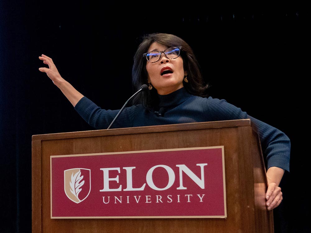 Wendy Suzuki confidently addresses the audience from behind a podium on stage during a presentation at Elon University.