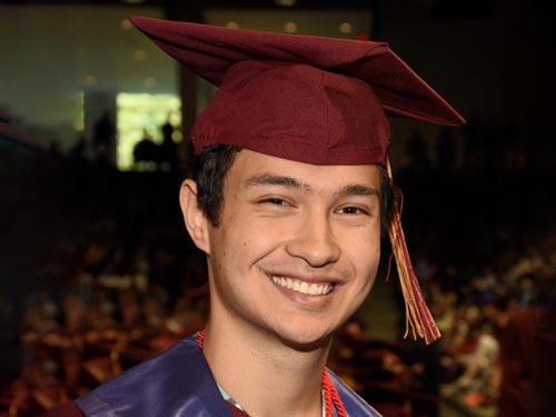 Nicholas Hom, adorned in a maroon graduation cap and gown, smiles proudly for the camera at Elon University.