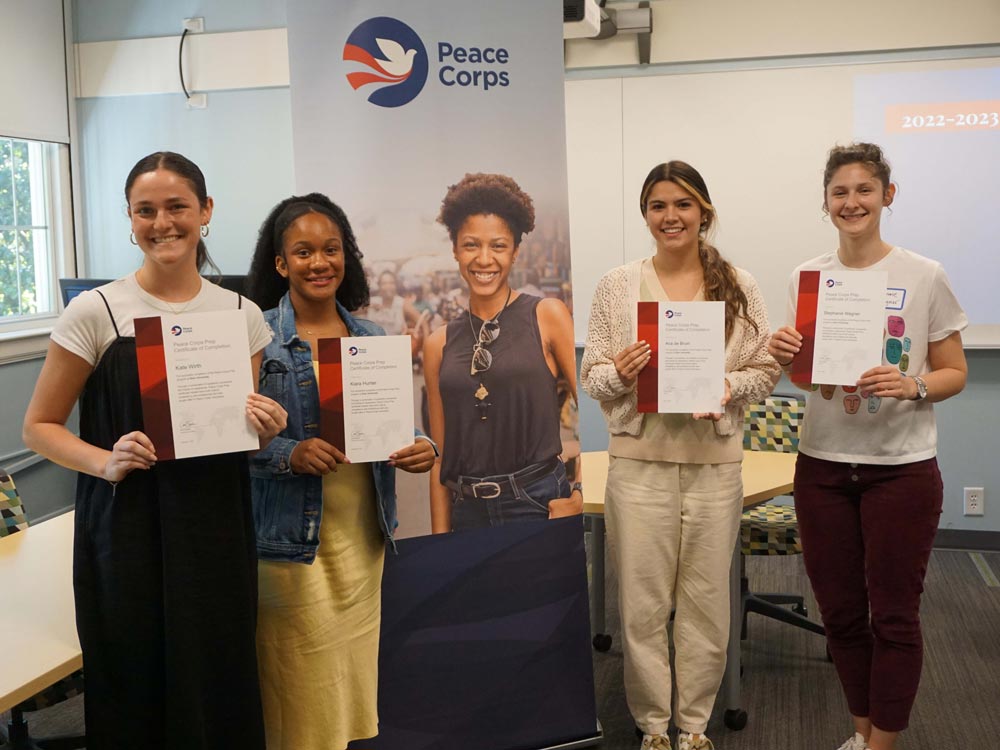 Standing beside a Peace Corps banner, two students on each side proudly display their completion certificates, celebrating their successful completion of the Peace Corps Prep program.
