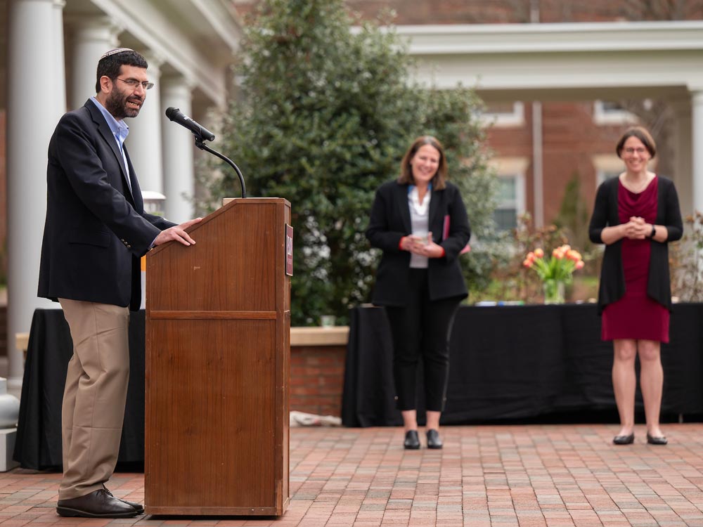 Professor of Religious Studies Geoffrey Claussen stands at a podium outdoors on Elon University's campus during an event celebrating the first decade of the Jewish Studies program.