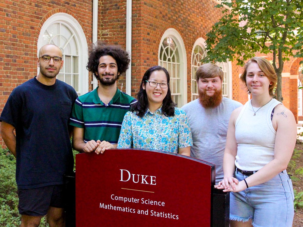 Hwayeon Ryu, accompanied by two students on each side, stands proudly behind the sign for Elon University's Duke Building, home to the Computer Science and Mathematics & Statistics departments.