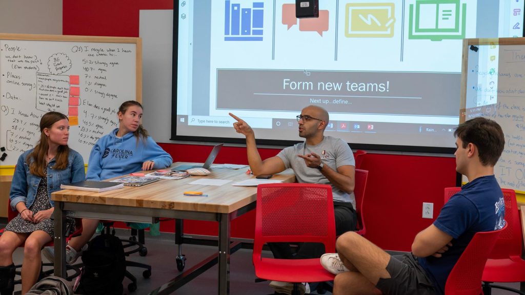 Four students gather around a wooden table, seated in vibrant red chairs, engaging in a discussion. Behind them, a whiteboard covered in notes and a digital presentation screen provide additional context.