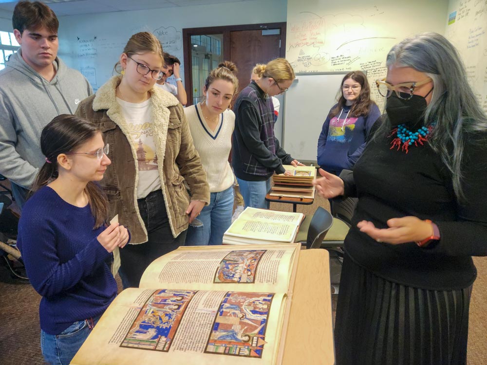 Professor of Art History Evan Gatti, conducting a guest lecture within an Art History class, gestures towards a detailed facsimile of a medieval European manuscript, while several students attentively observe.