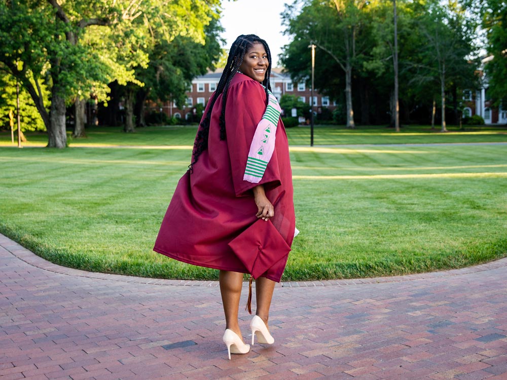 Arianne Payne, elegantly dressed in her maroon graduation gown, holding her cap, walks away from the camera with a smile, while looking back, on a brick walkway at Elon's campus.