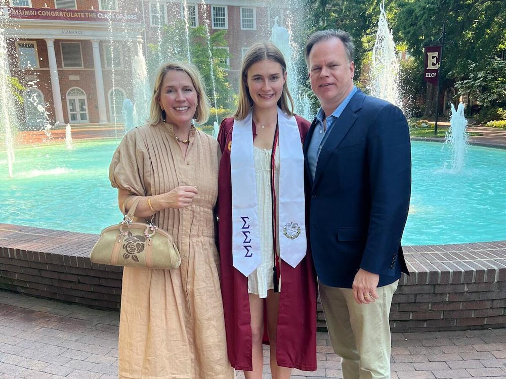 Clay and Cameron Smith, along with their daughter Margaret in her graduation gown, photographed in front of Elon University's Fonville Fountain.