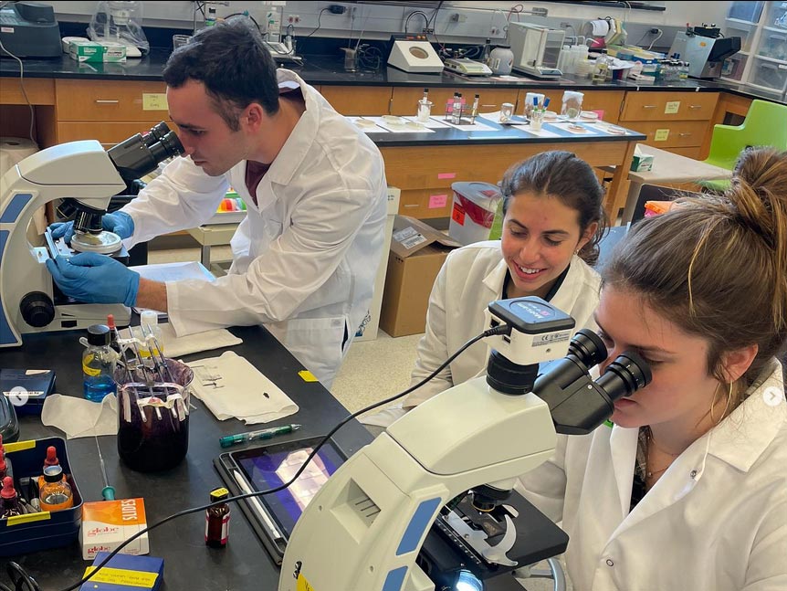 Three students in lab coats, two peering into microscopes, work diligently in Elon's Biology lab.