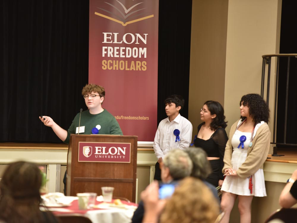An Elon Freedom Scholar stands confidently at a podium, delivering a presentation during the symposium. Three fellow scholars stand beside the podium, with a prominent banner displaying the Freedom Scholars logo in the background.