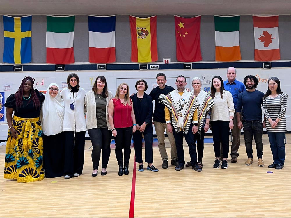 Seven faculty members from the Department of World Languages and Cultures engage with the local community in the Marvin B. Smith Elementary School gym, with various international flags hanging on the wall in the background.