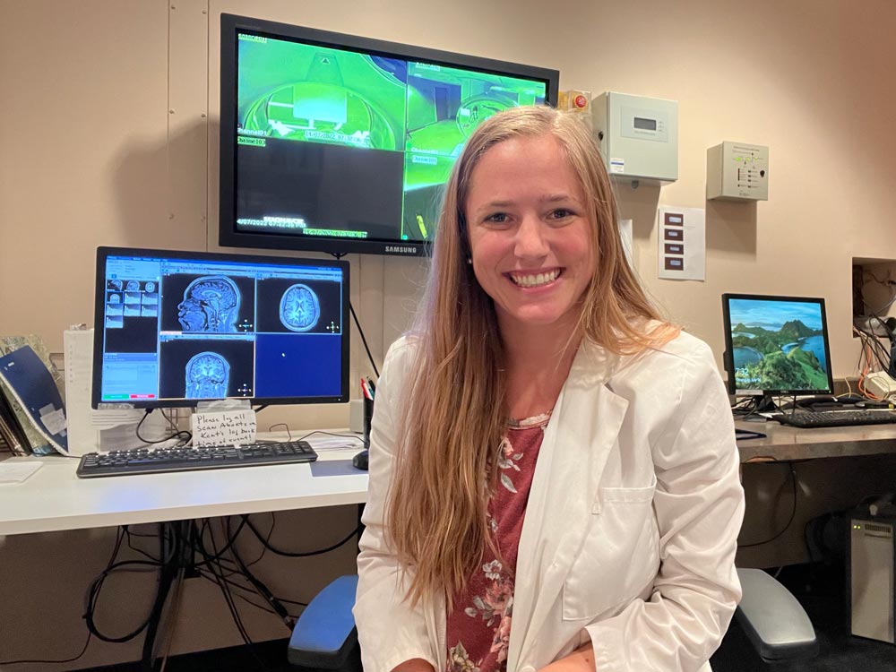 Kathleen Hupfeld, donning a lab coat, smiles for the camera while seated in front of computer monitors displaying MRI images.