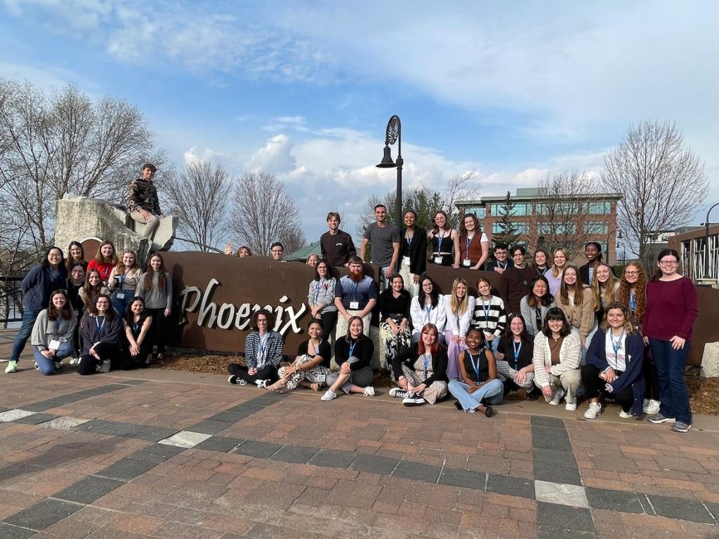 A substantial gathering of Elon students and faculty assembles around a monument bearing the word 'Phoenix' while on a visit to Eau Claire, Wisconsin, for the National Conference on Undergraduate Research.