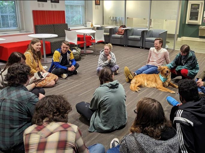 A group of students sits on the floor in a common area, gathered for a coffee gathering. One student sits beside a friendly golden retriever, giving them affectionate head pats.