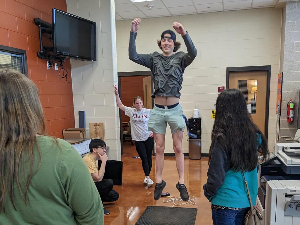 During National Biomechanics Day, a high school student jumps to test their airtime, with other students observing the experiment.