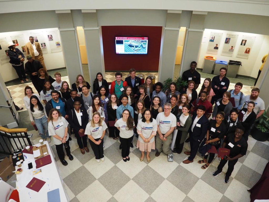 A group of student and faculty attendees of the Integrating Research in Science conference come together for a photograph in the lobby of Elon University's McMichael building.