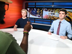 Two students talk with a mentor while sitting at a TV news desk.