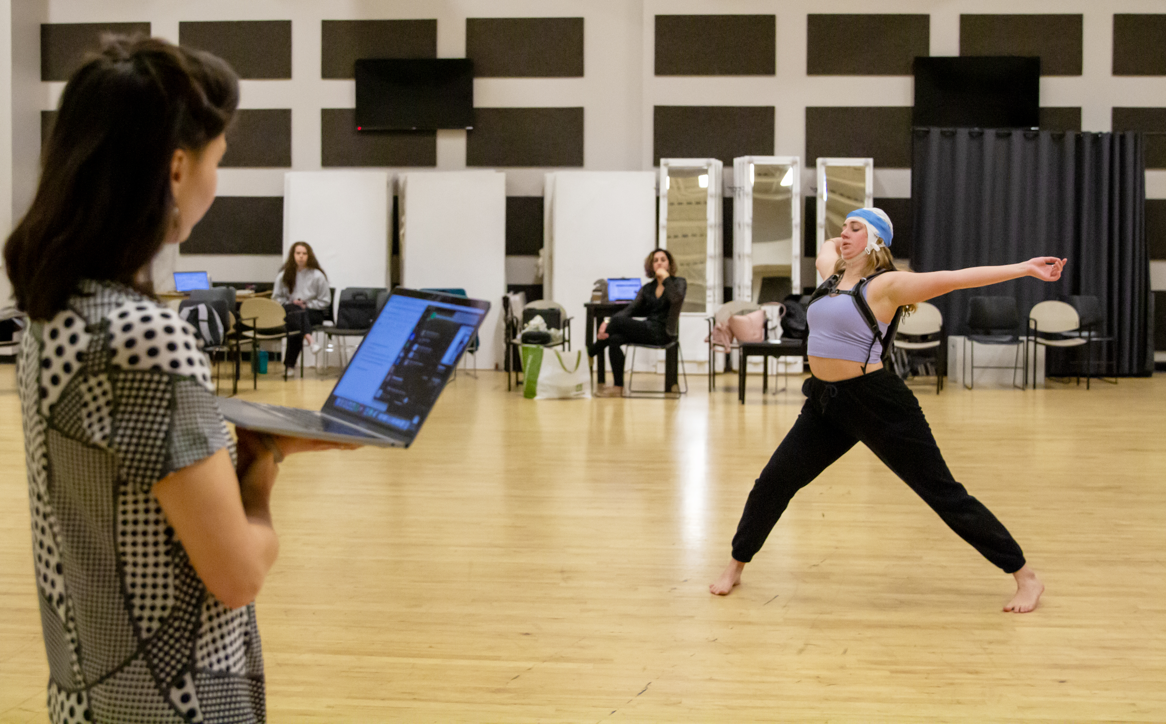 a female dancer wears an EEG cap while dancing in a large rehearsal space. A female professor observes while holding her laptop.