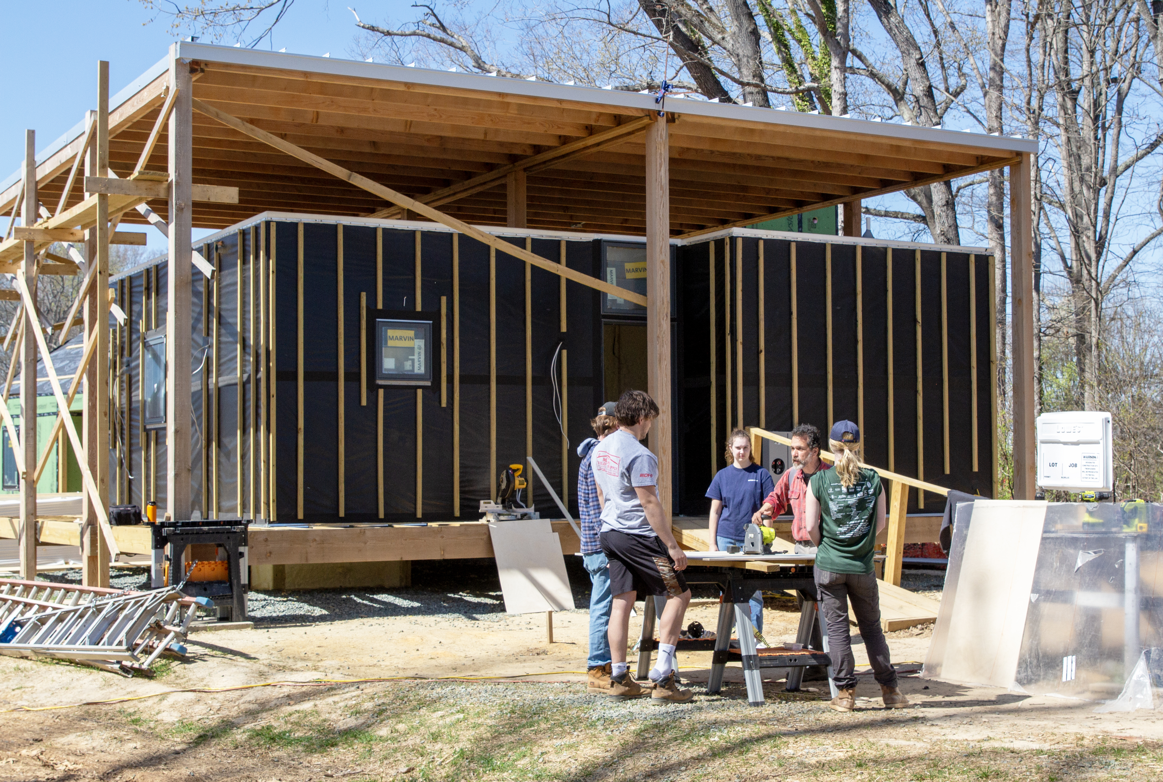 A group of students and a professor around a table saw in front of a small house under construction